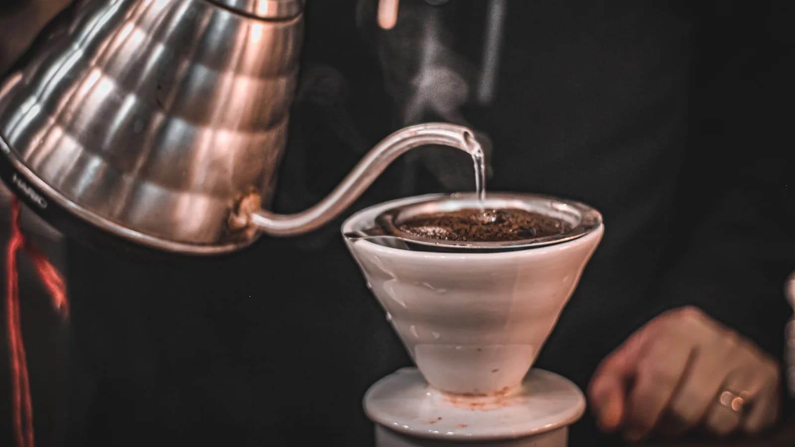 A gooseneck kettle pouring hot water over coffee grounds in a white ceramic cone dripper, steam rising.